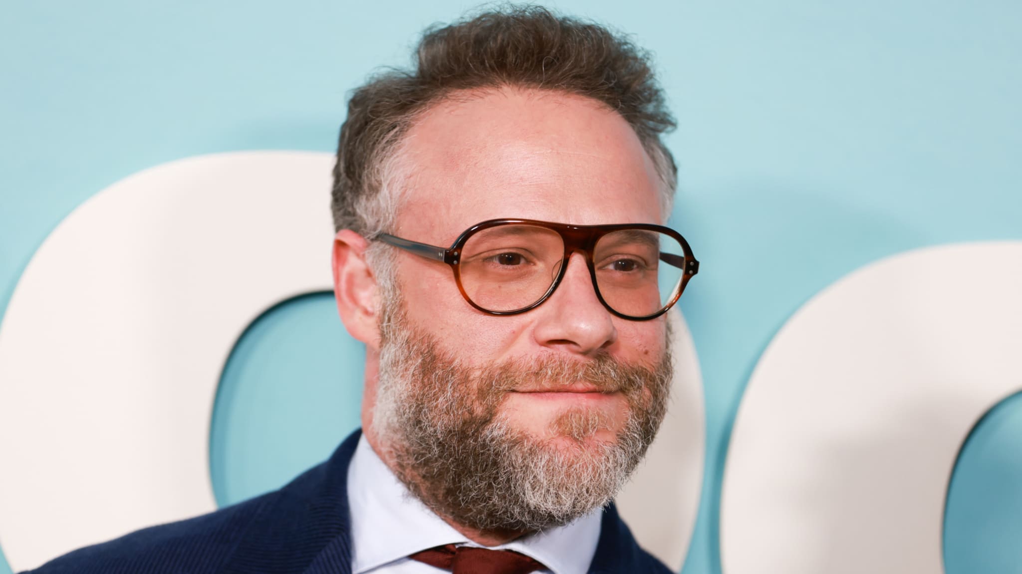 Seth Rogen with glasses and a beard is wearing a suit and tie, standing against a light blue background with white letters.