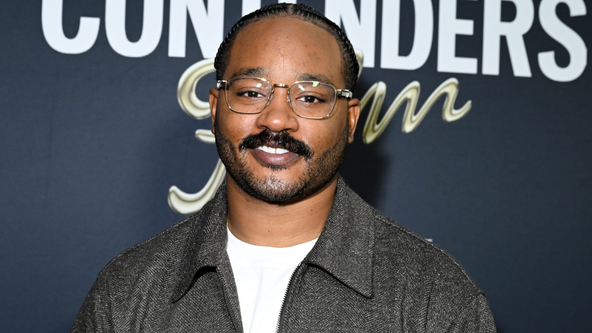 Ryan Coogler with glasses and a mustache, wearing a gray jacket, stands in front of a backdrop with the word "CONTENDERS" visible.