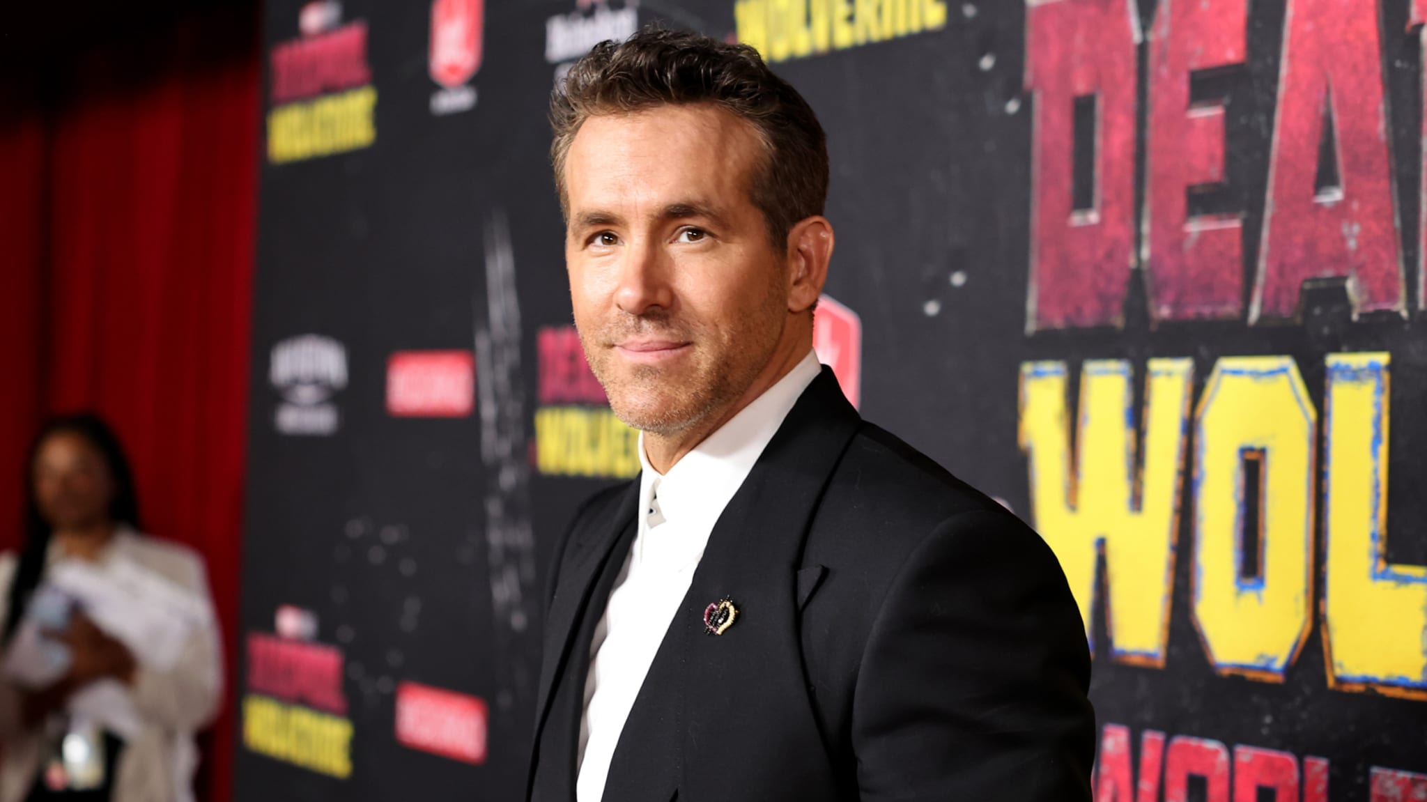 Ryan Reynolds at a red carpet event, wearing a black suit and white shirt, with "Deadpool" and "Wolverine" signage in the background.