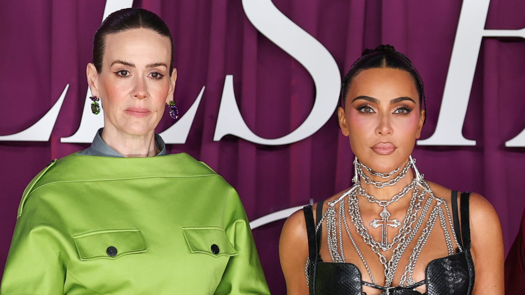 Sarah Paulson in a green outfit and Kim Kardashian in a black dress with layered necklaces at an event with a purple backdrop.
