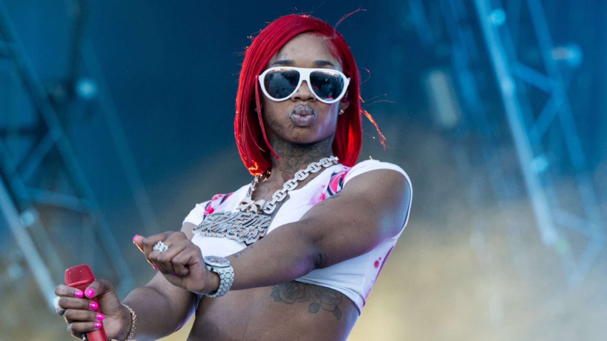 Sexyy Red with red hair and sunglasses, holding a microphone on stage, wearing a white top and chain necklace.
