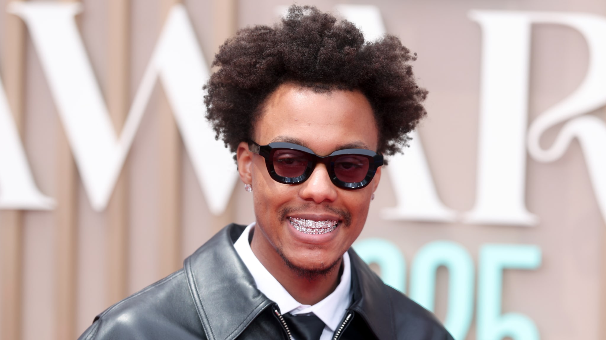 Skilla Baby with curly hair, wearing sunglasses and braces, smiles at an event. They are dressed in a leather jacket and tie.