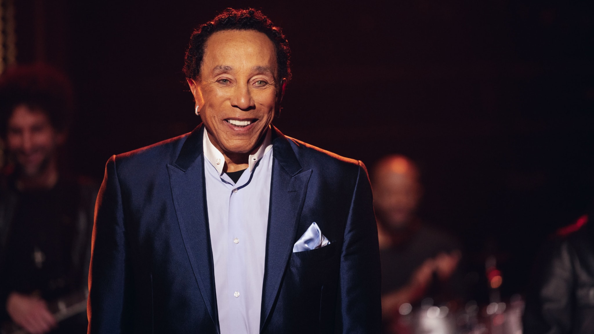 Smokey Robinson in in a blue suit and white shirt smiles on stage with musicians in the background.