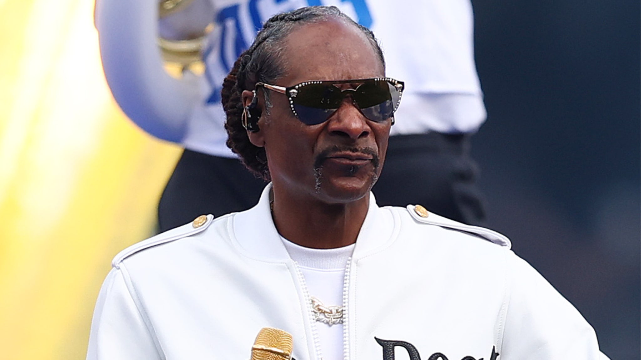 Snoop Dogg wearing sunglasses and a white jacket, holding a gold microphone.