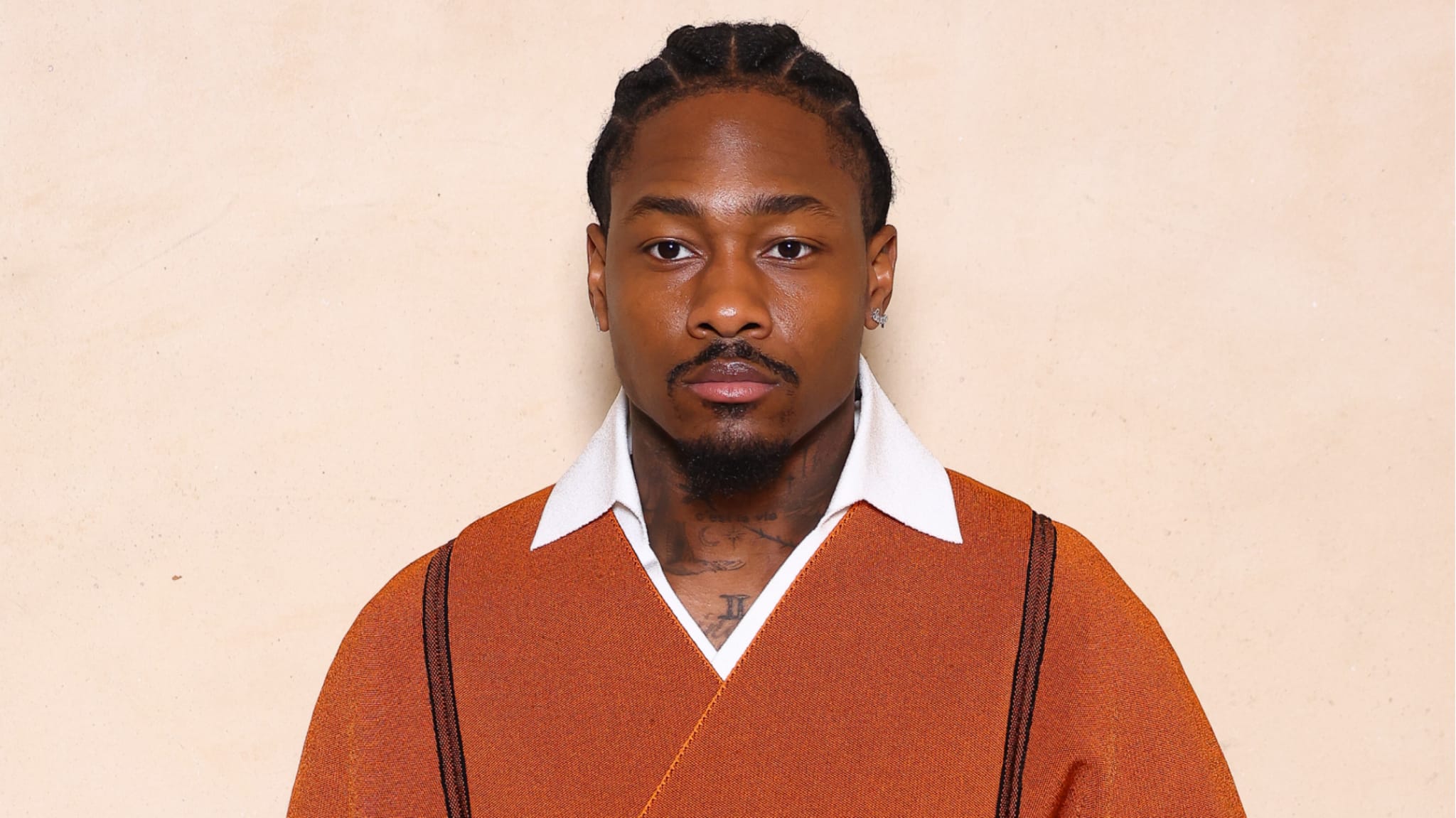 A man with braided hair wearing an orange outfit with a white collar, posing against a neutral background.