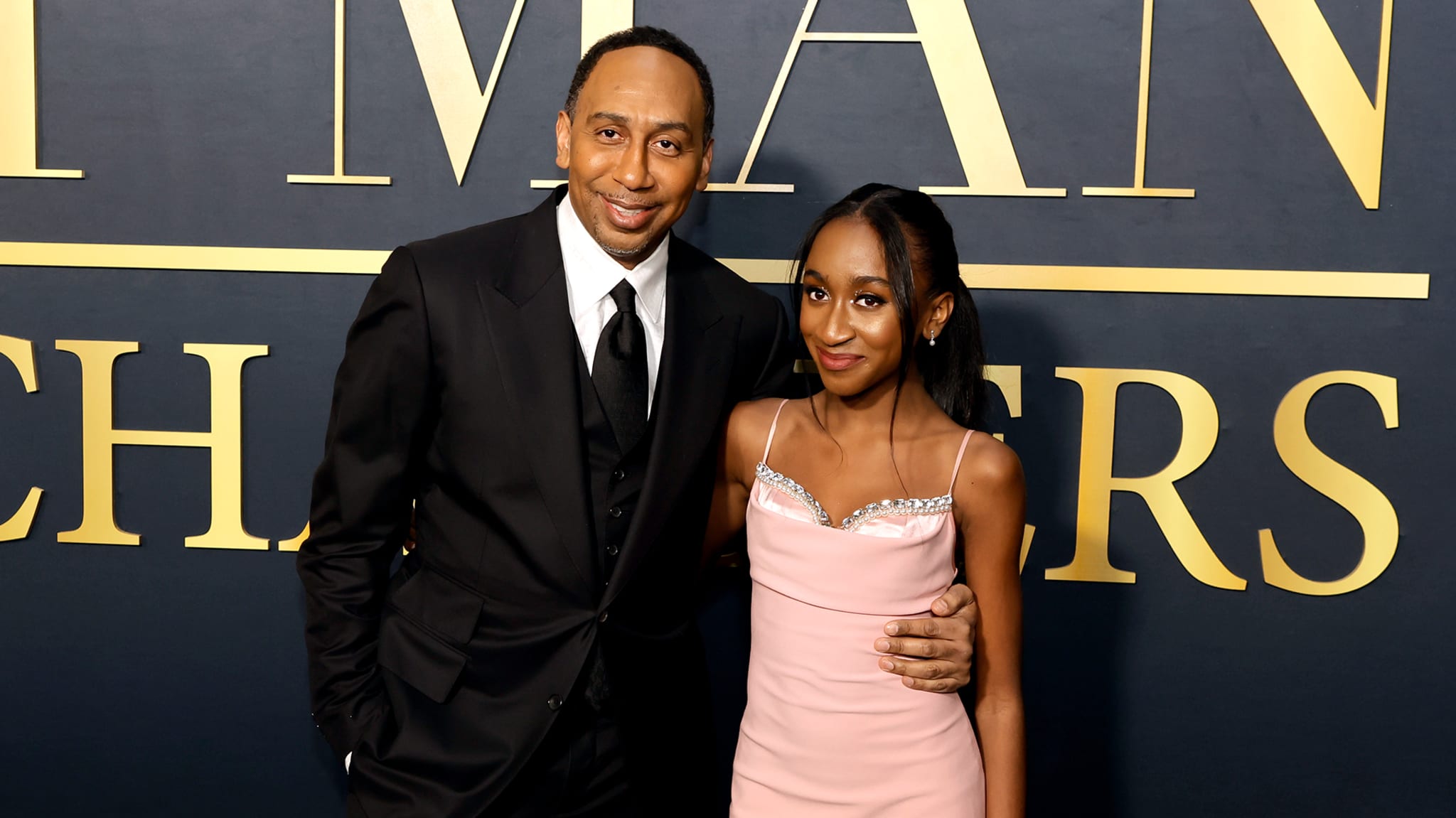 A man in a black suit and a young woman in a pink dress pose together at an event with a dark backdrop featuring gold lettering.