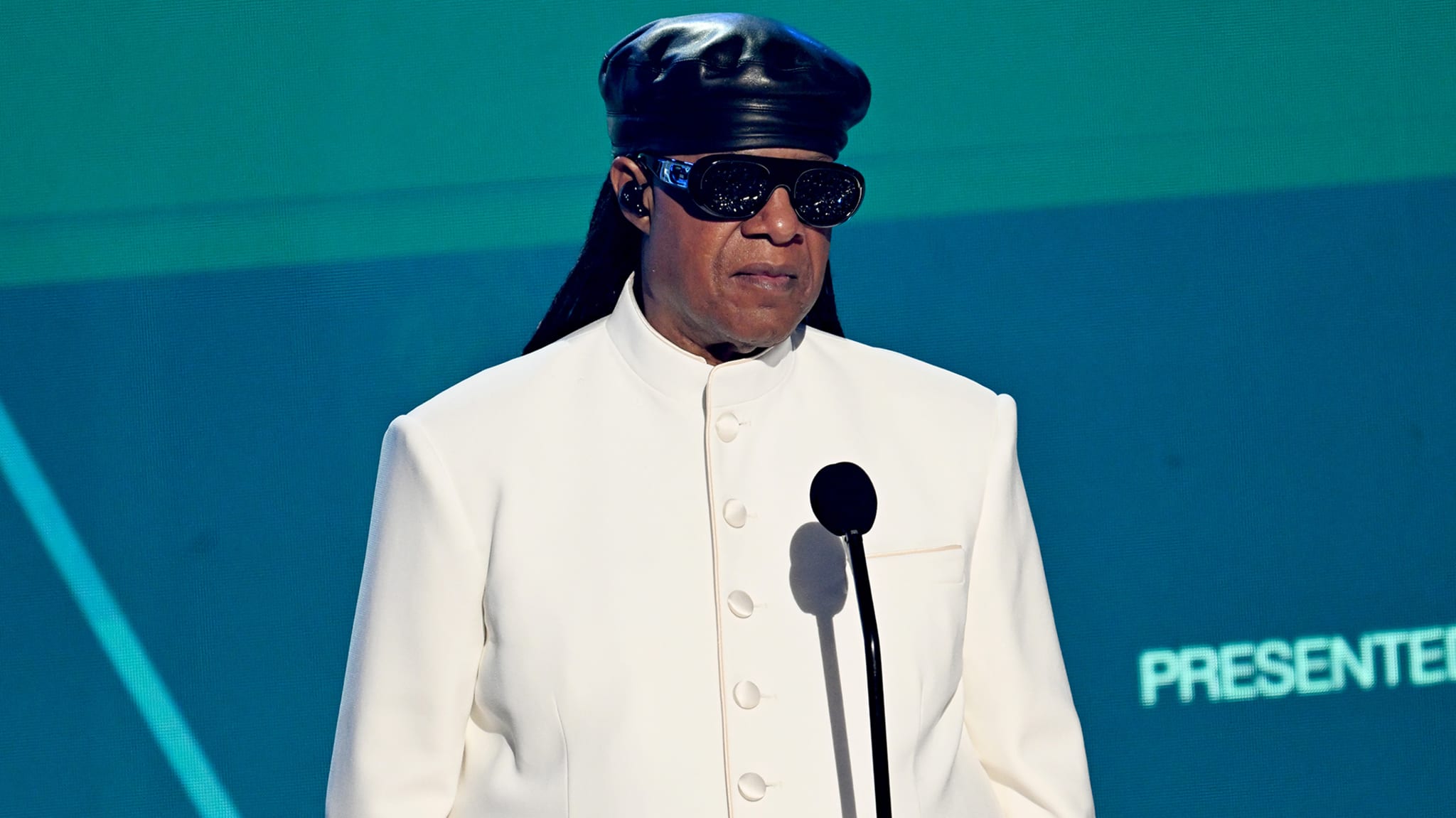 Stevie Wonder in a white suit and black hat, wearing sunglasses, stands at a microphone on stage.