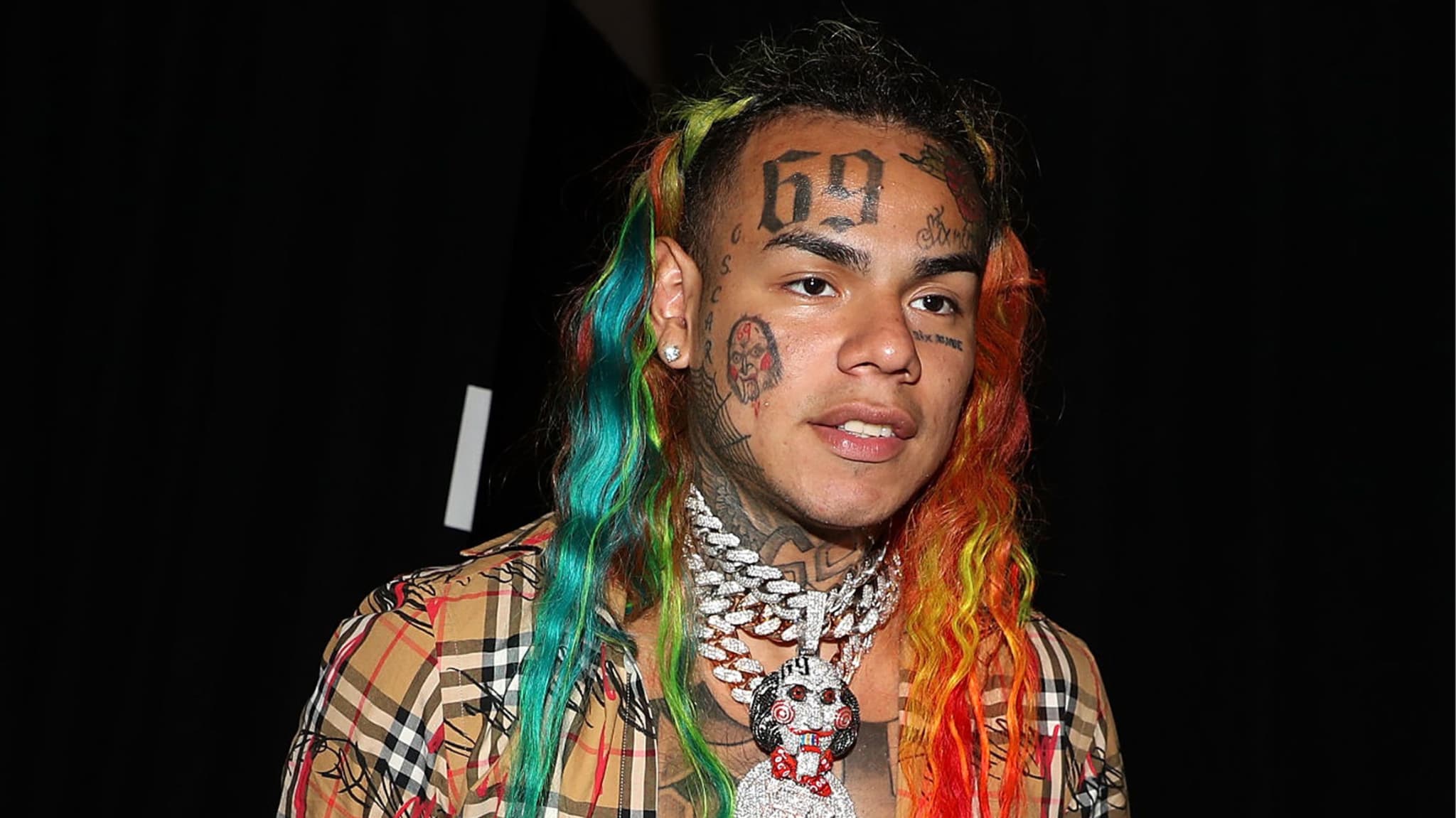 Rapper Tekashi 6ix9ine with rainbow hair, face tattoos, and a plaid shirt, wearing a large diamond necklace.