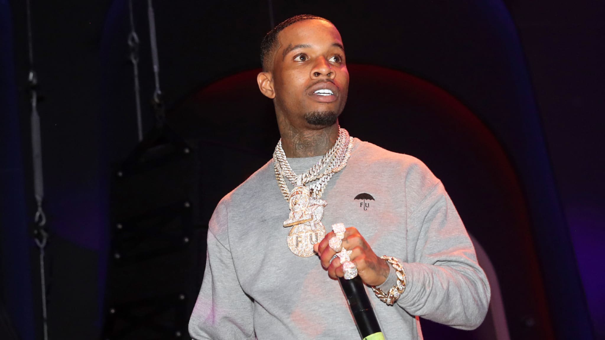 Tory Lanez in a gray sweatshirt holds a microphone on stage, wearing multiple chains and rings. The background is dark with stage lights.