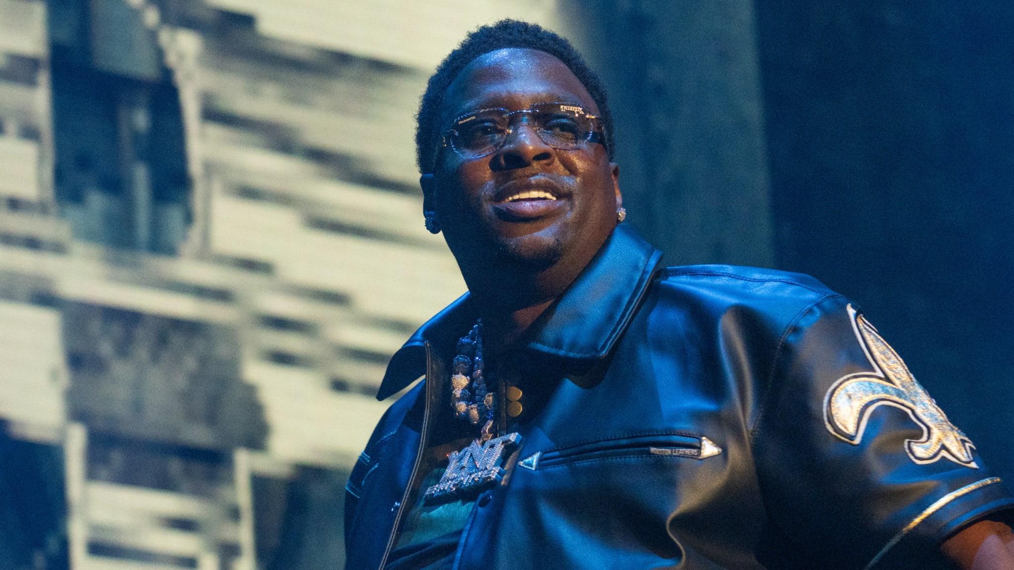 A man wearing glasses and a black leather jacket with a fleur-de-lis emblem, performing on stage.