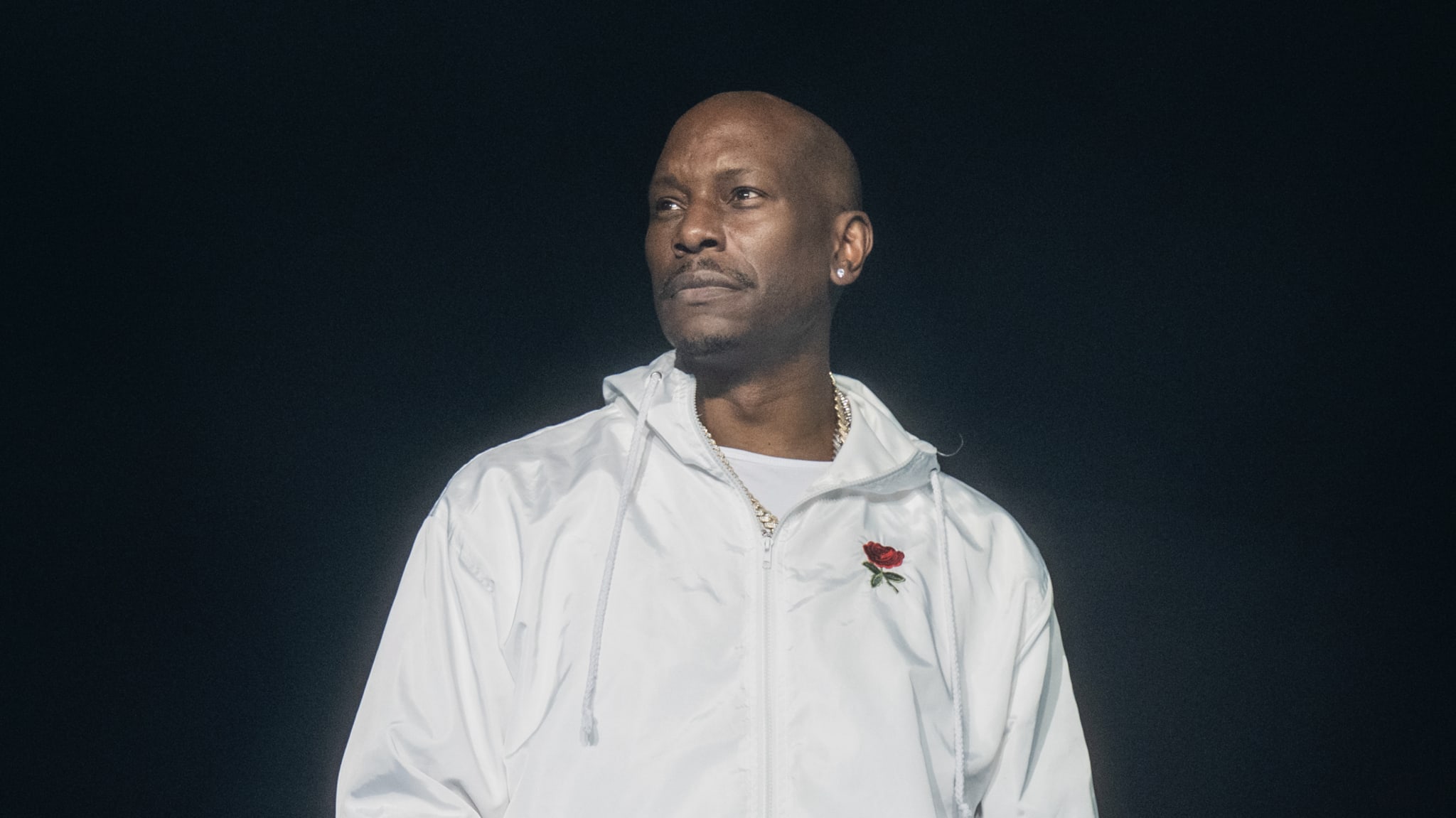 Tyrese in a white jacket with a rose emblem stands against a dark background.