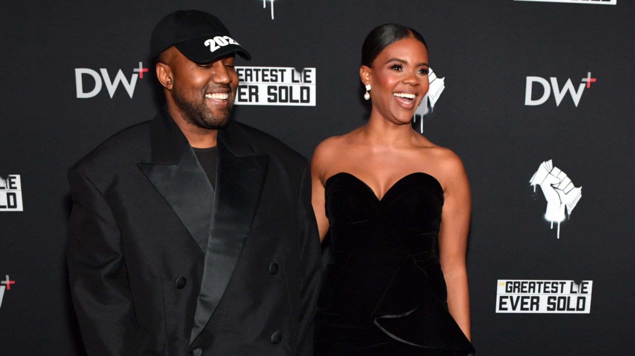 Kanye West and Candace Owens smiling at an event, with a black backdrop featuring logos and text.