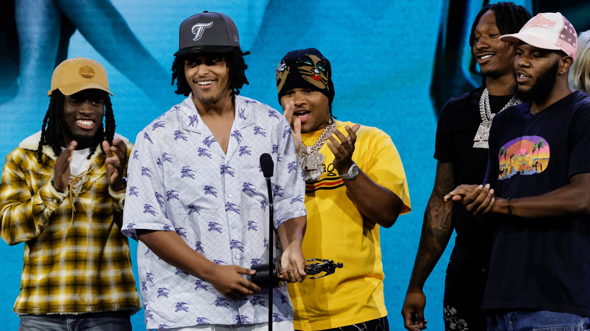 A group of five men on stage, one holding a trophy. They are wearing casual clothing and hats, smiling and clapping.