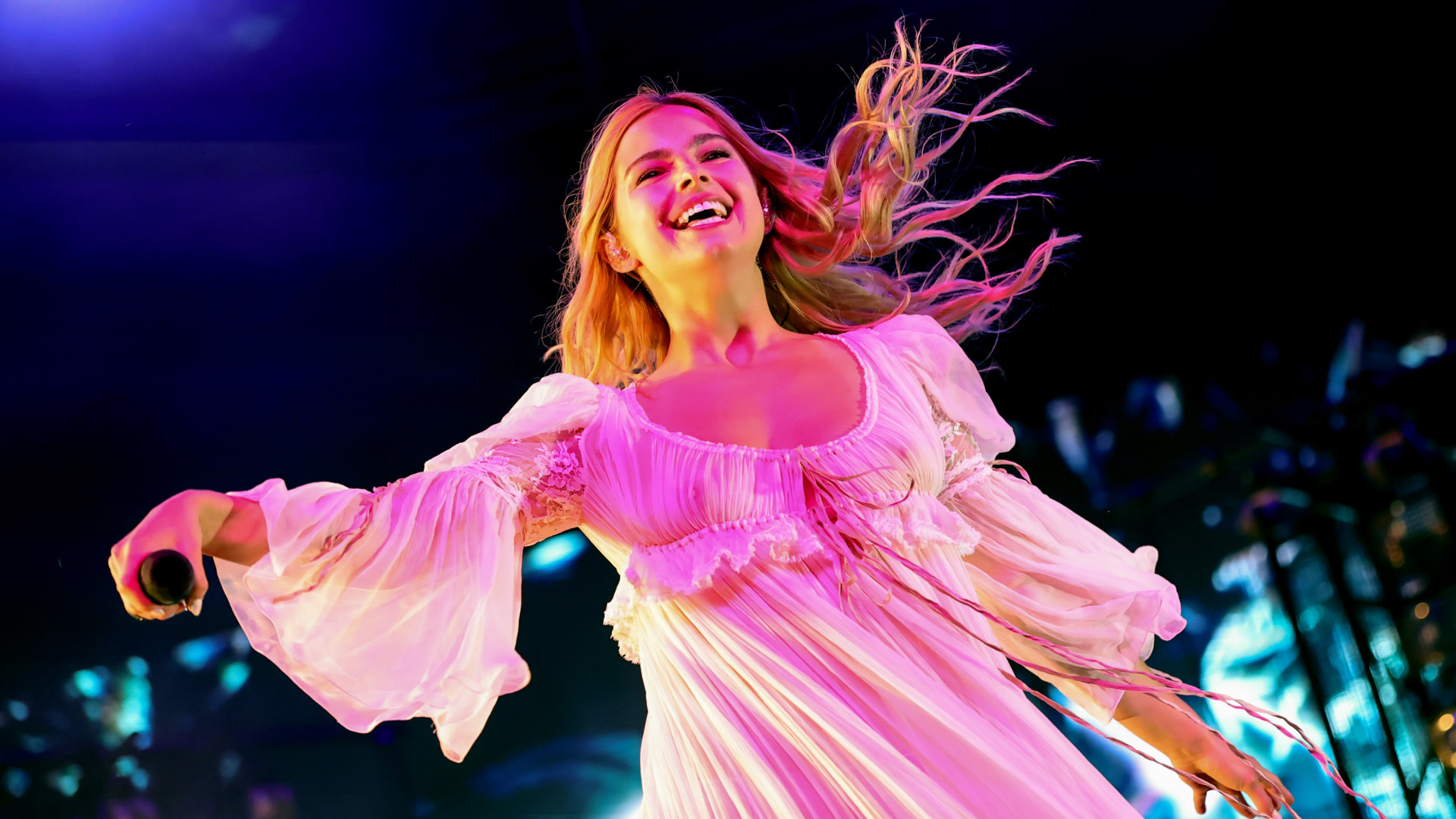 Addison Rae in a flowing white dress performs on stage, smiling and holding a microphone, with colorful lights in the background.