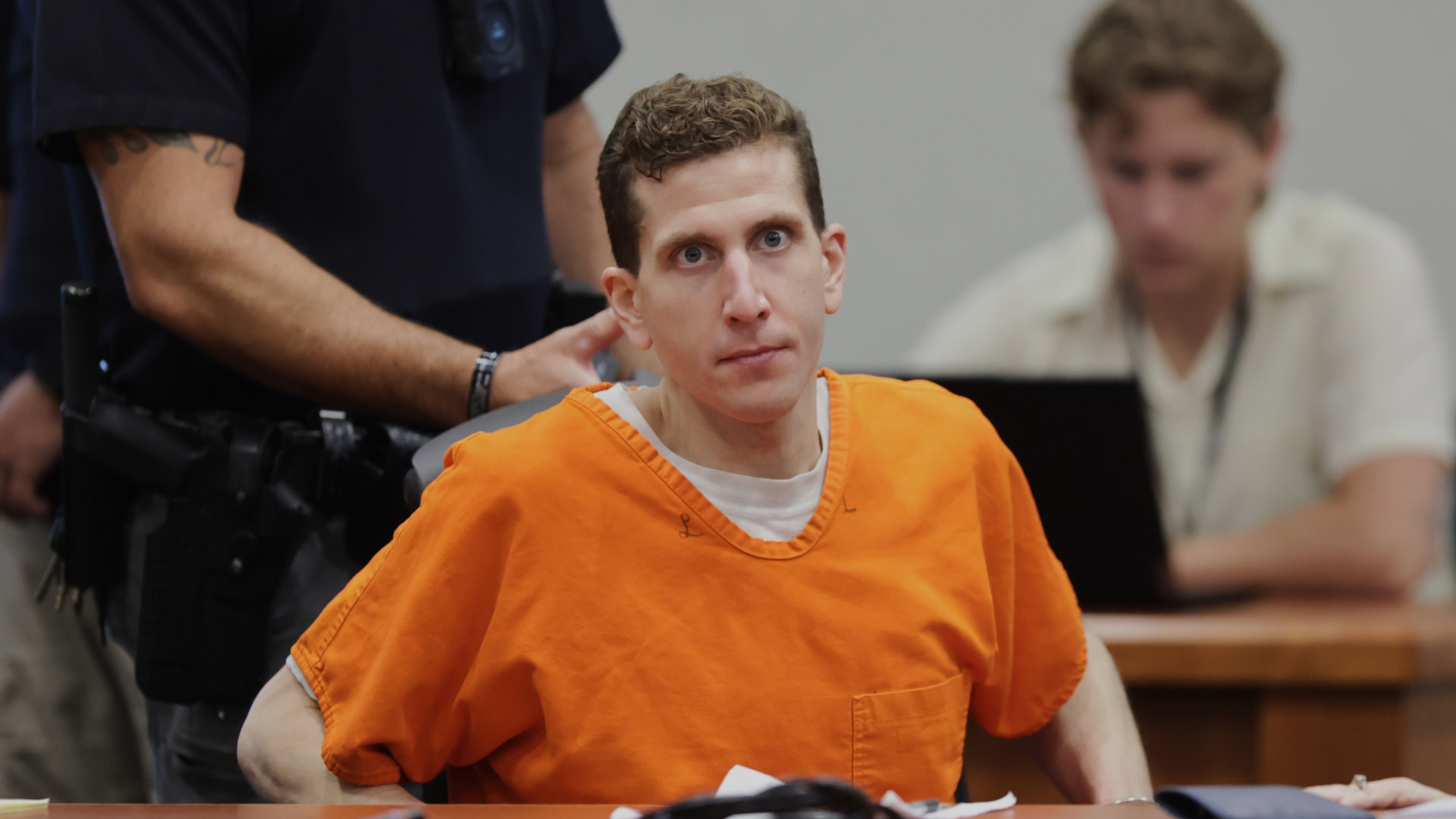 A man in an orange prison jumpsuit sits in a courtroom, with a guard behind him and another person in the background.