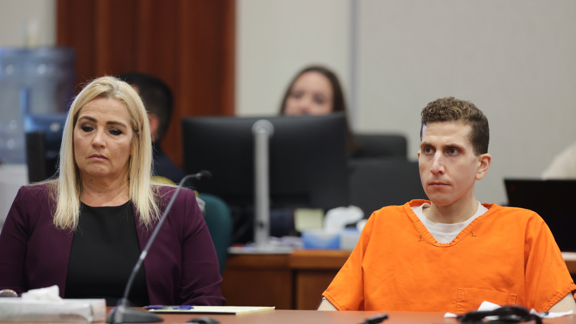 A man in an orange jumpsuit sits beside a woman in a courtroom. Both appear serious, surrounded by legal documents and microphones.