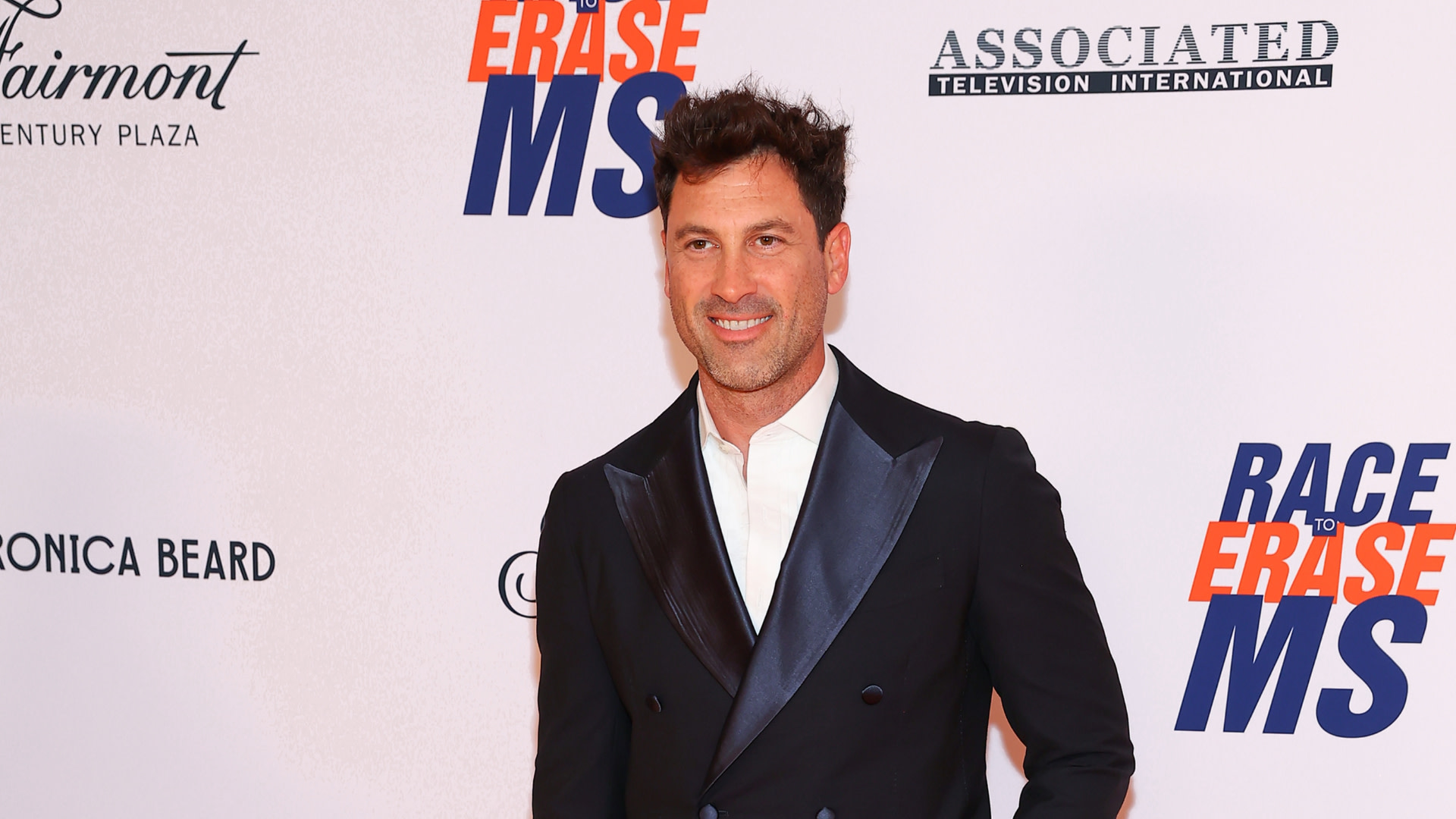 A man in a dark suit and white shirt stands smiling at the "Race to Erase MS" event, with a logo backdrop.