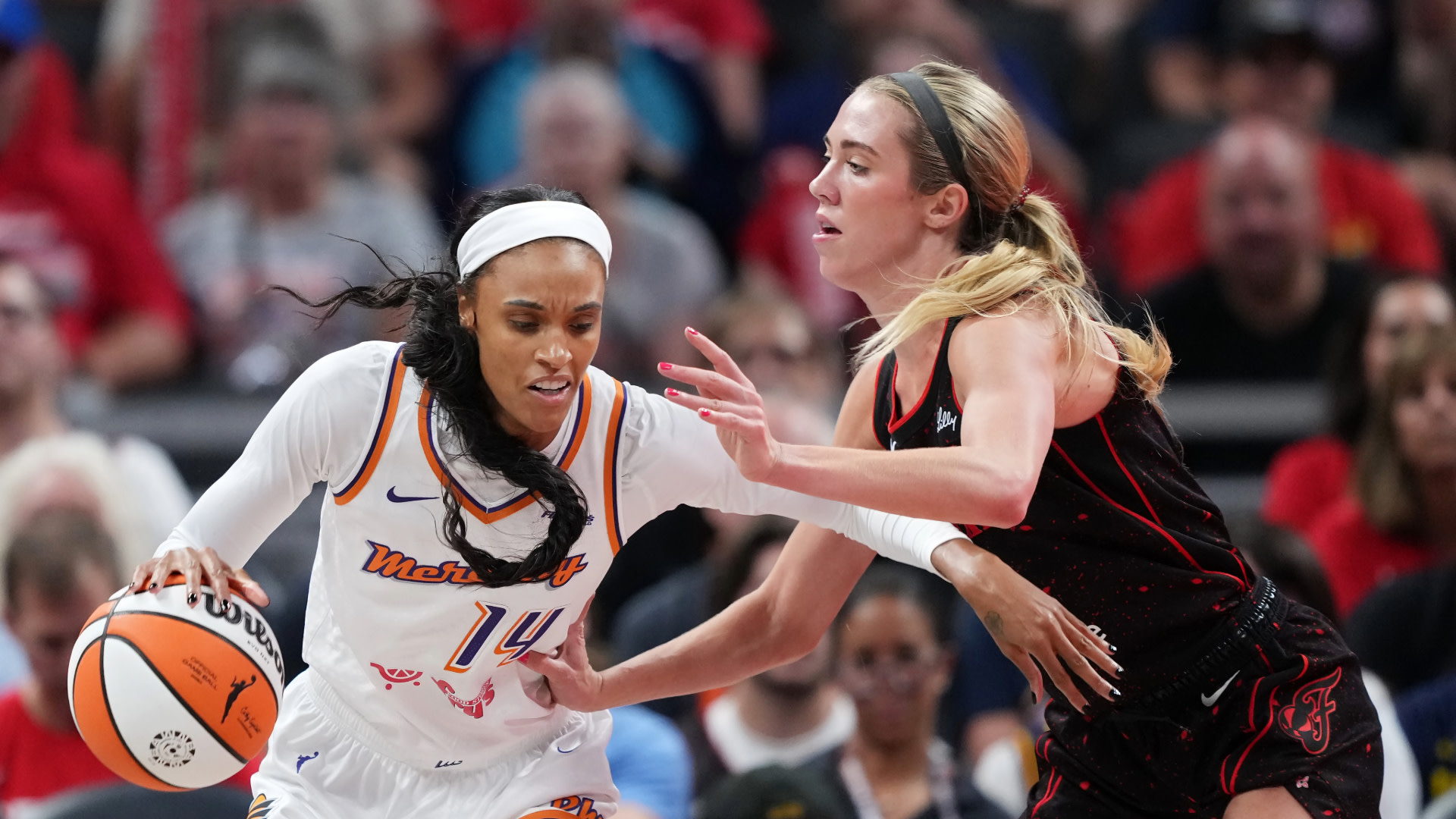 Two female basketball players in action on the court, one in a white jersey and the other in a black jersey, with a crowd in the background.