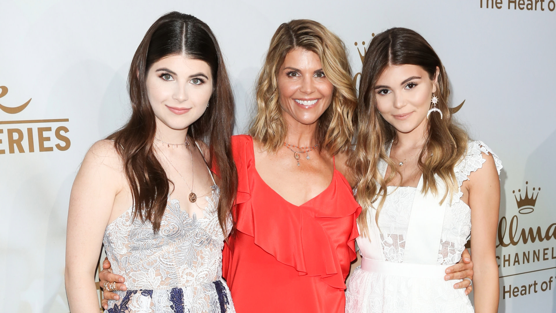 Three women posing at an event: Lori Loughlin in a red dress with her daughters, Isabella and Olivia Jade, in white dresses.