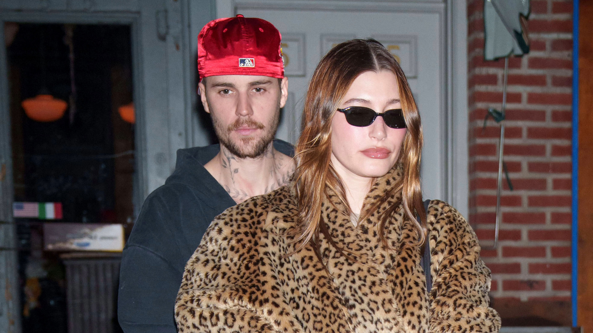 Justin Bieber in a red cap and Hailey Bieber in a leopard print coat and sunglasses, standing outside a building.