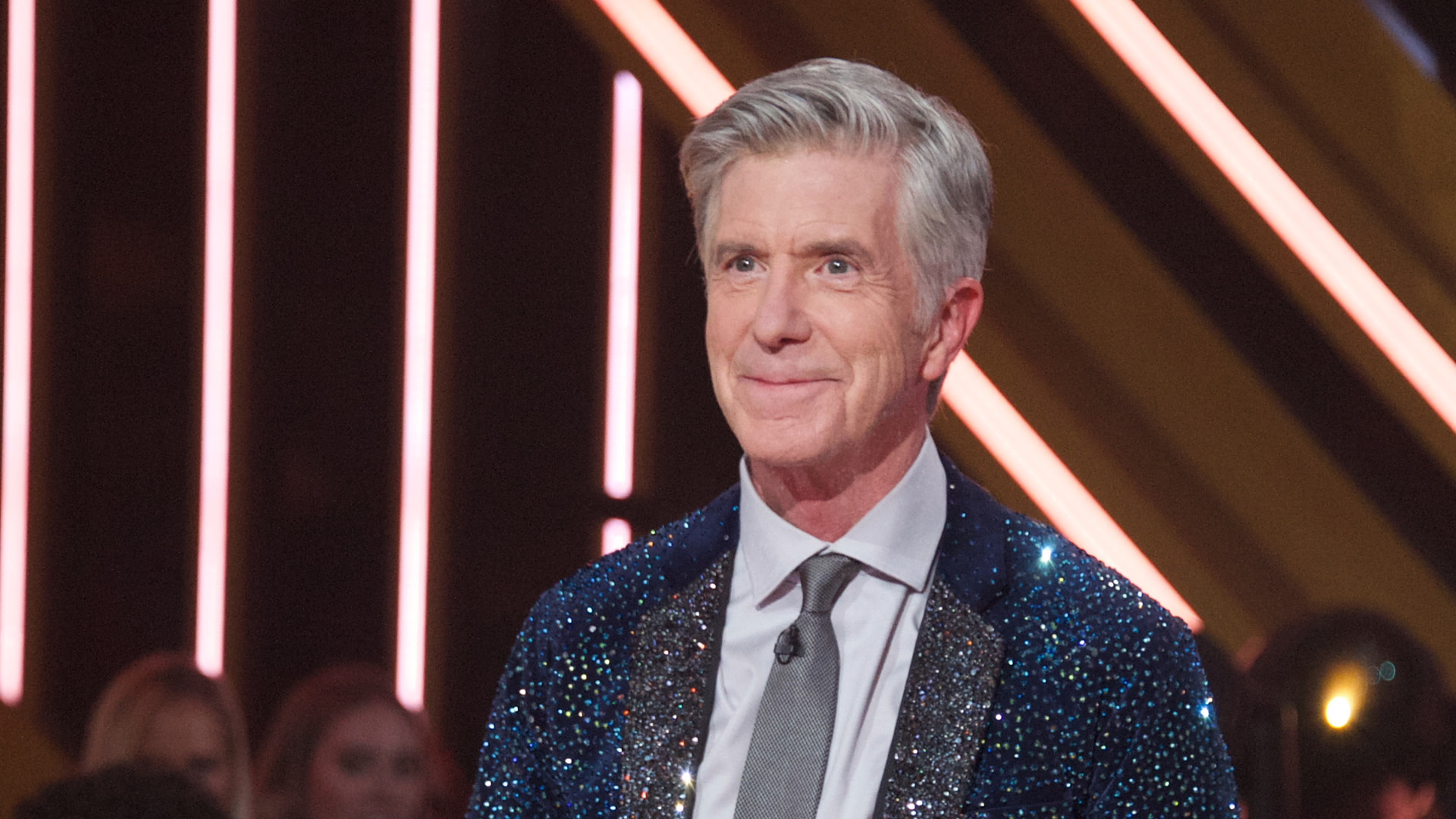 A man with gray hair wearing a sparkly jacket and tie stands in front of a lit background.