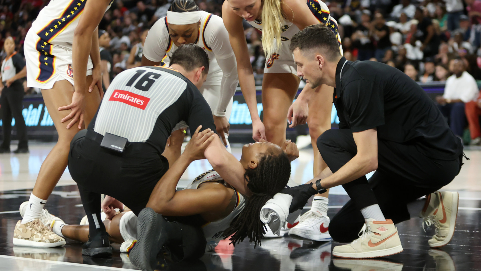 A basketball player is on the court, surrounded by teammates and officials, receiving assistance. The scene is tense and concerned.