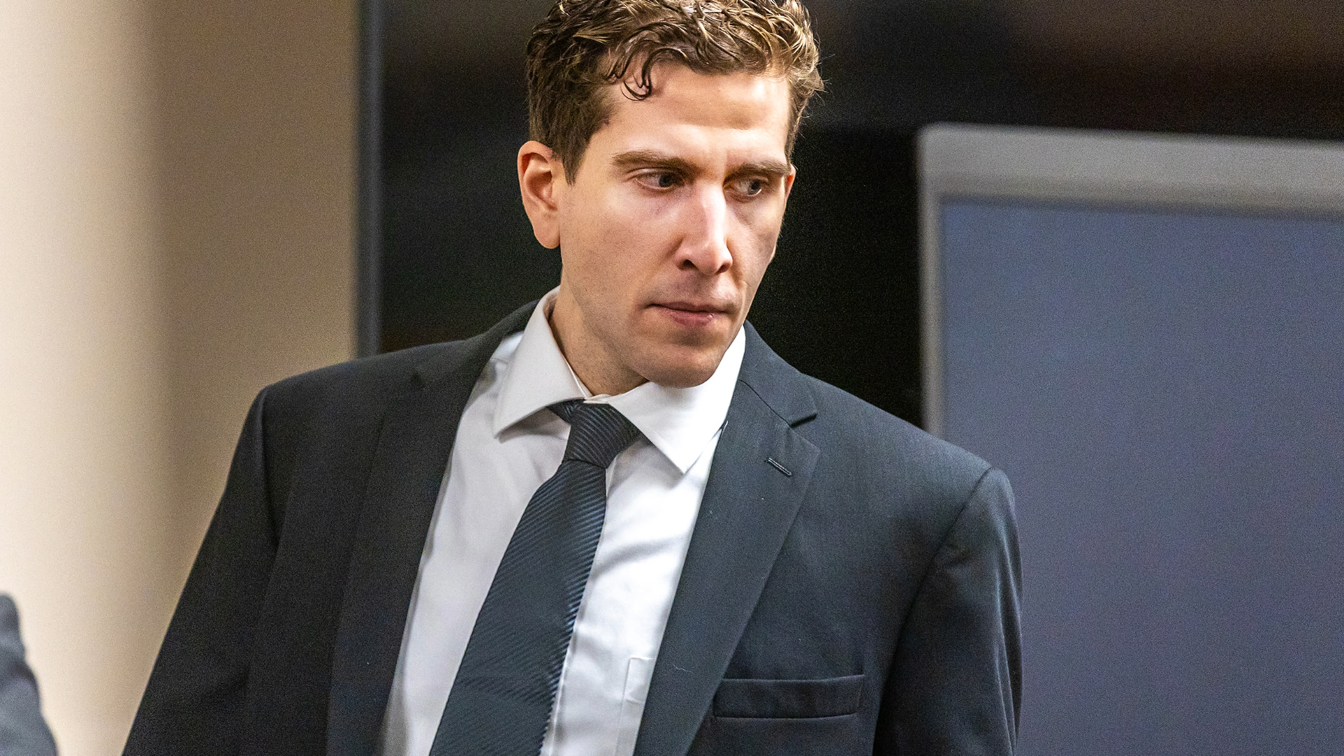 A man in a suit and tie stands indoors, looking to the side, with a serious expression.