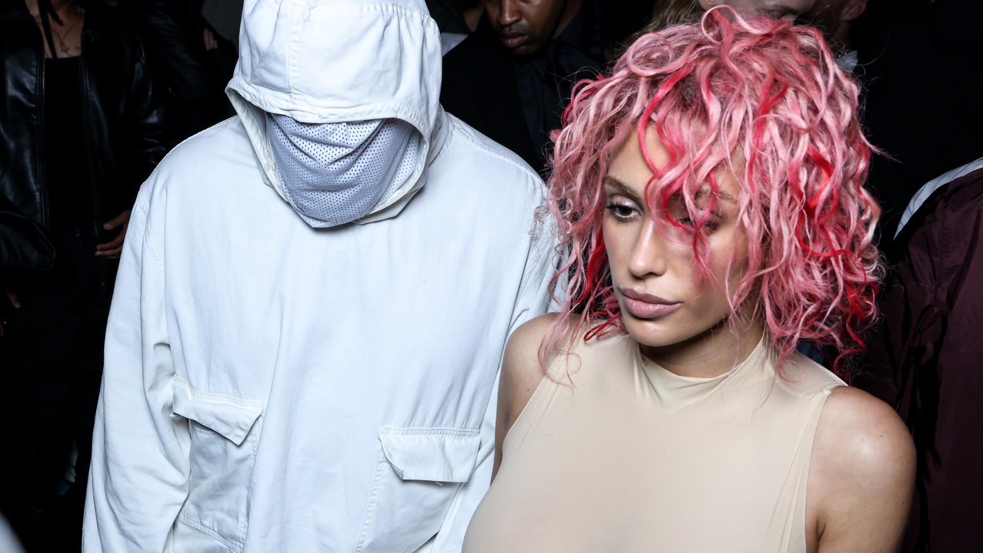 Ye in a white hooded outfit with face covered stands next to Bianca Censori with curly pink hair in a beige top.