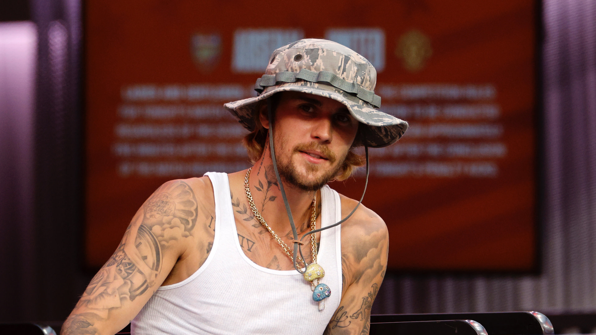 Justin Bieber wearing a camo bucket hat and white tank top, showcasing tattoos, with a blurred background.