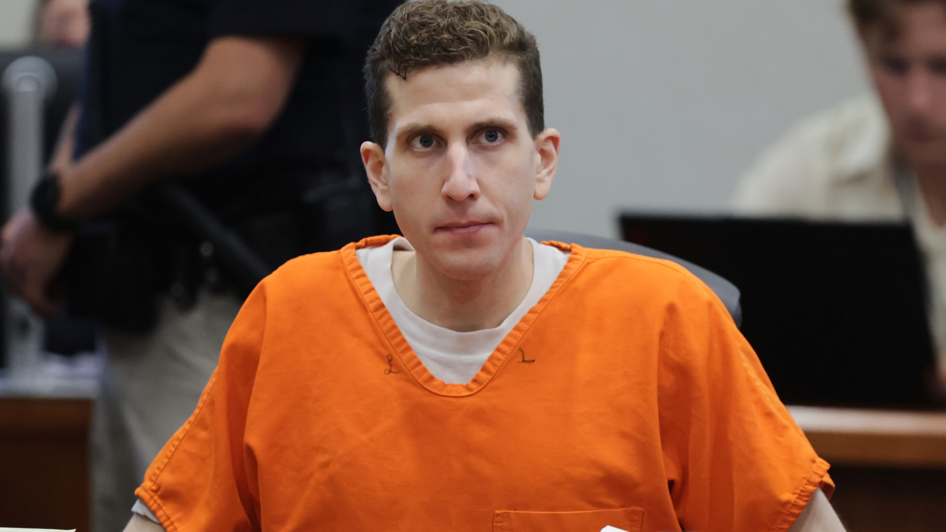A man in an orange prison jumpsuit sits in a courtroom, looking forward.