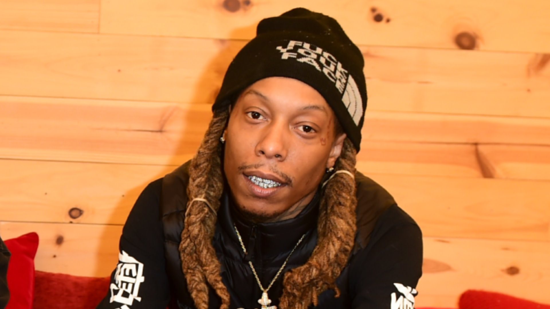 Rapper Chris King with long braids, wearing a black beanie and jacket, sitting indoors against a wooden wall.