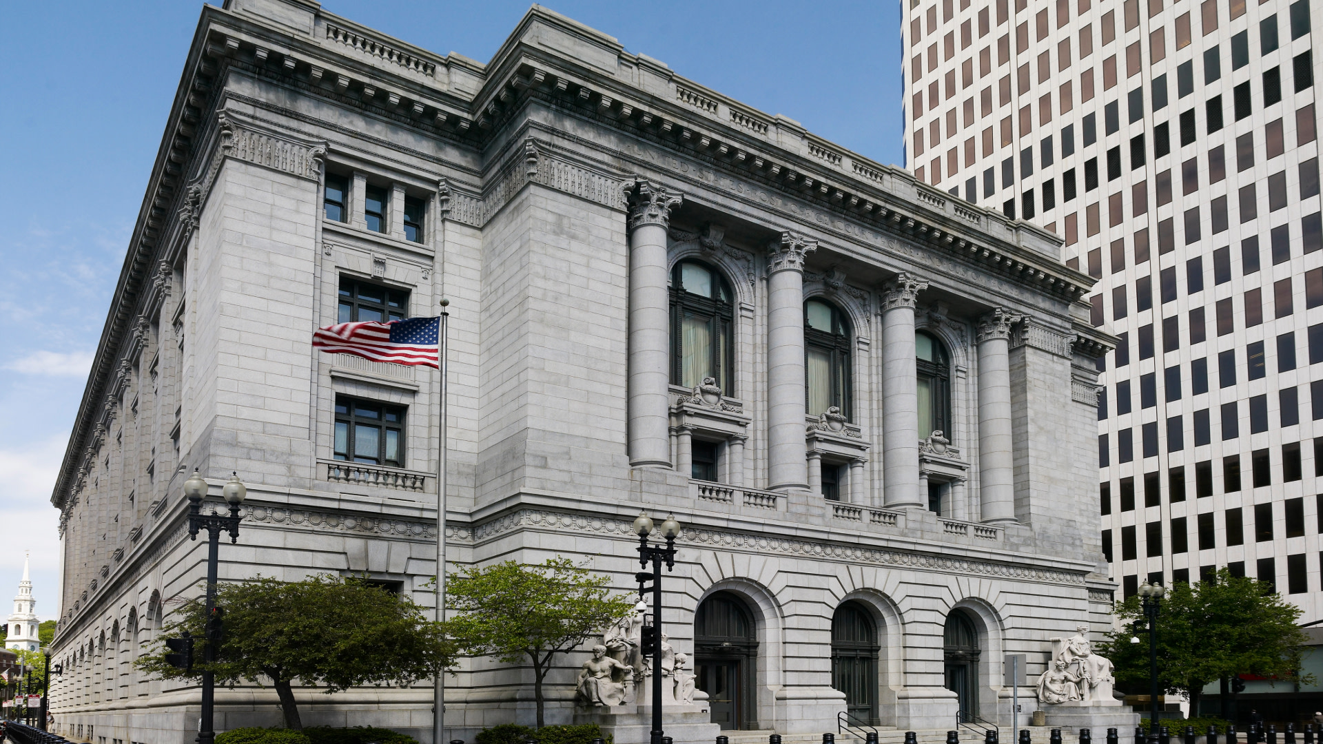 A historic stone building with columns and an American flag, set against a modern skyscraper.
