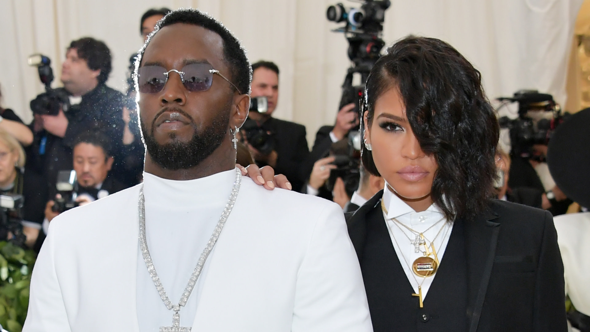 Sean "Diddy" Combs in a white suit and sunglasses with Cassie Ventura in a black suit, posing at an event.