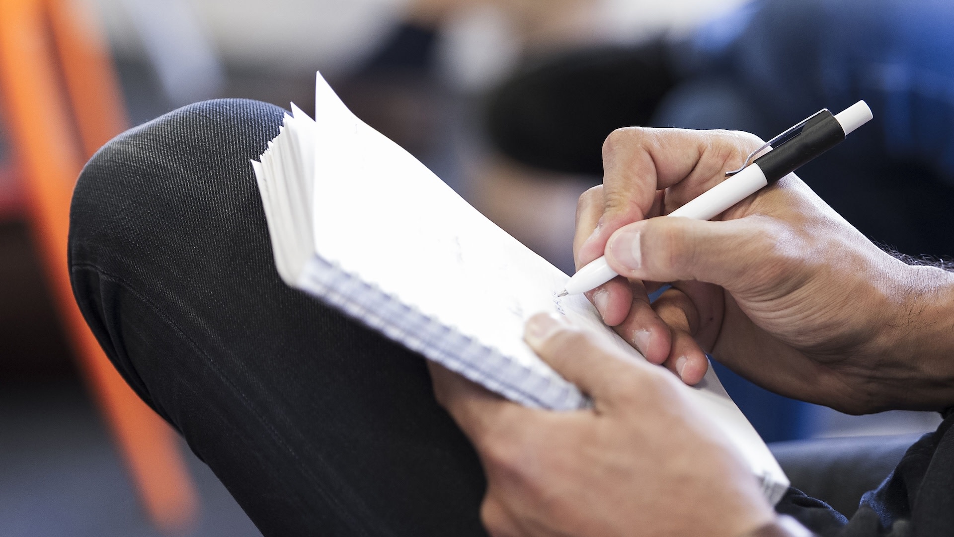 A person writing in a spiral notebook with a pen, seated and focused.