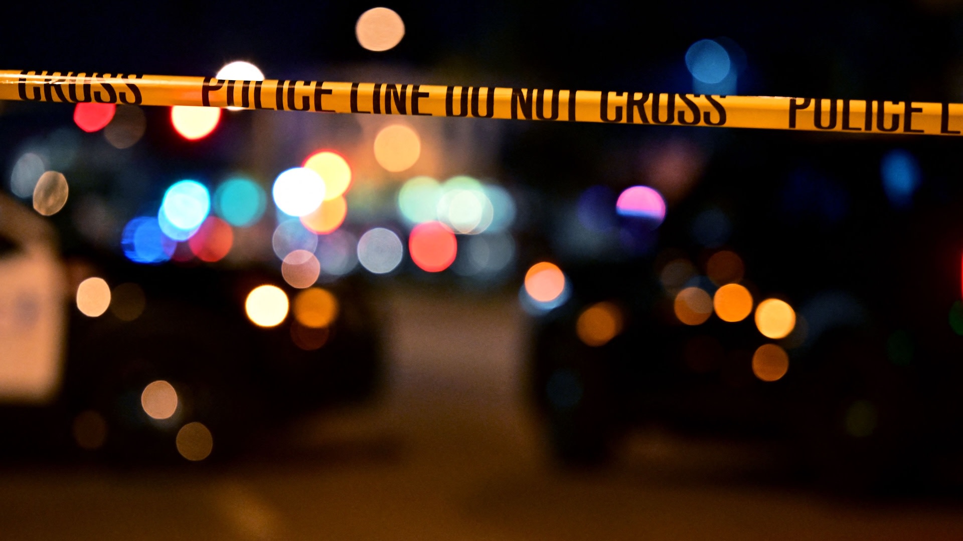 Police tape with "Do Not Cross" message, blurred lights and police cars in the background, creating a crime scene atmosphere.