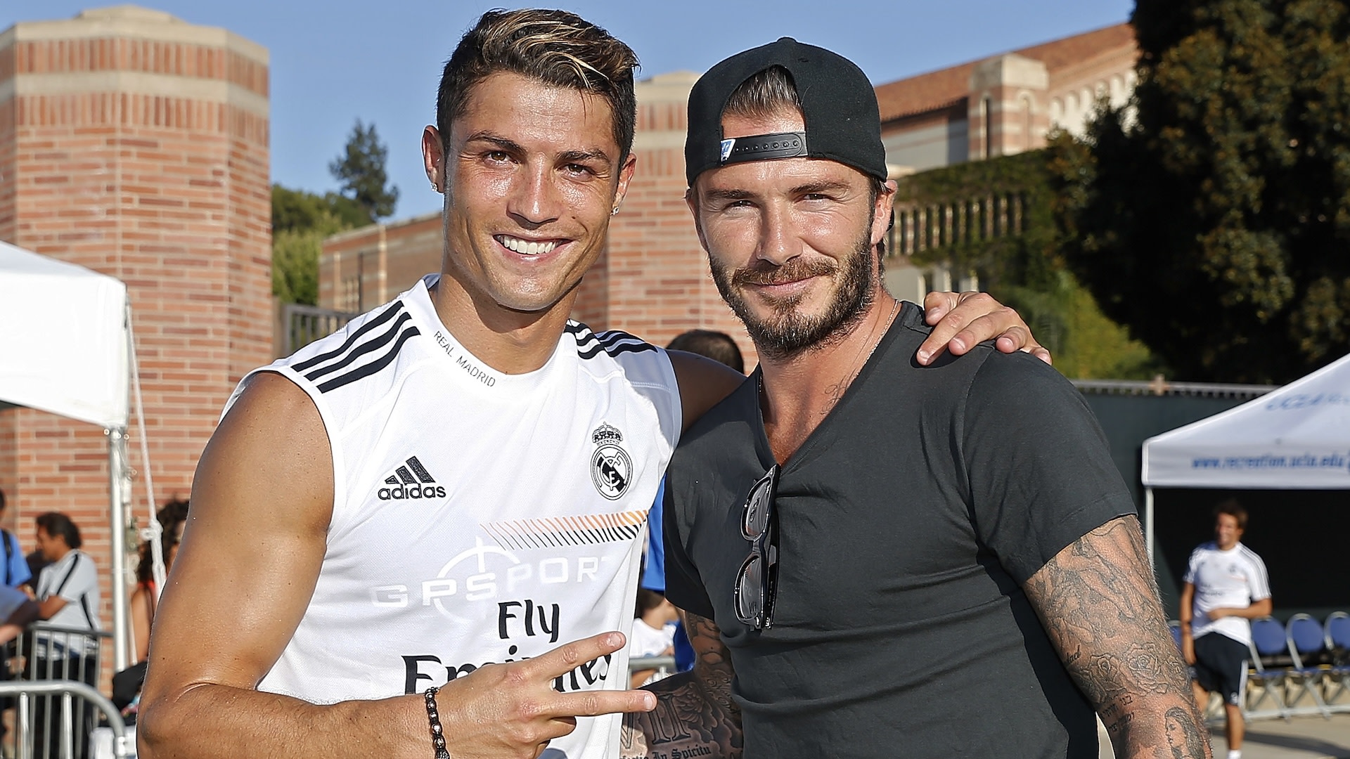 Cristiano Ronaldo in a Real Madrid training jersey poses with David Beckham, wearing a black t-shirt and cap, outdoors.