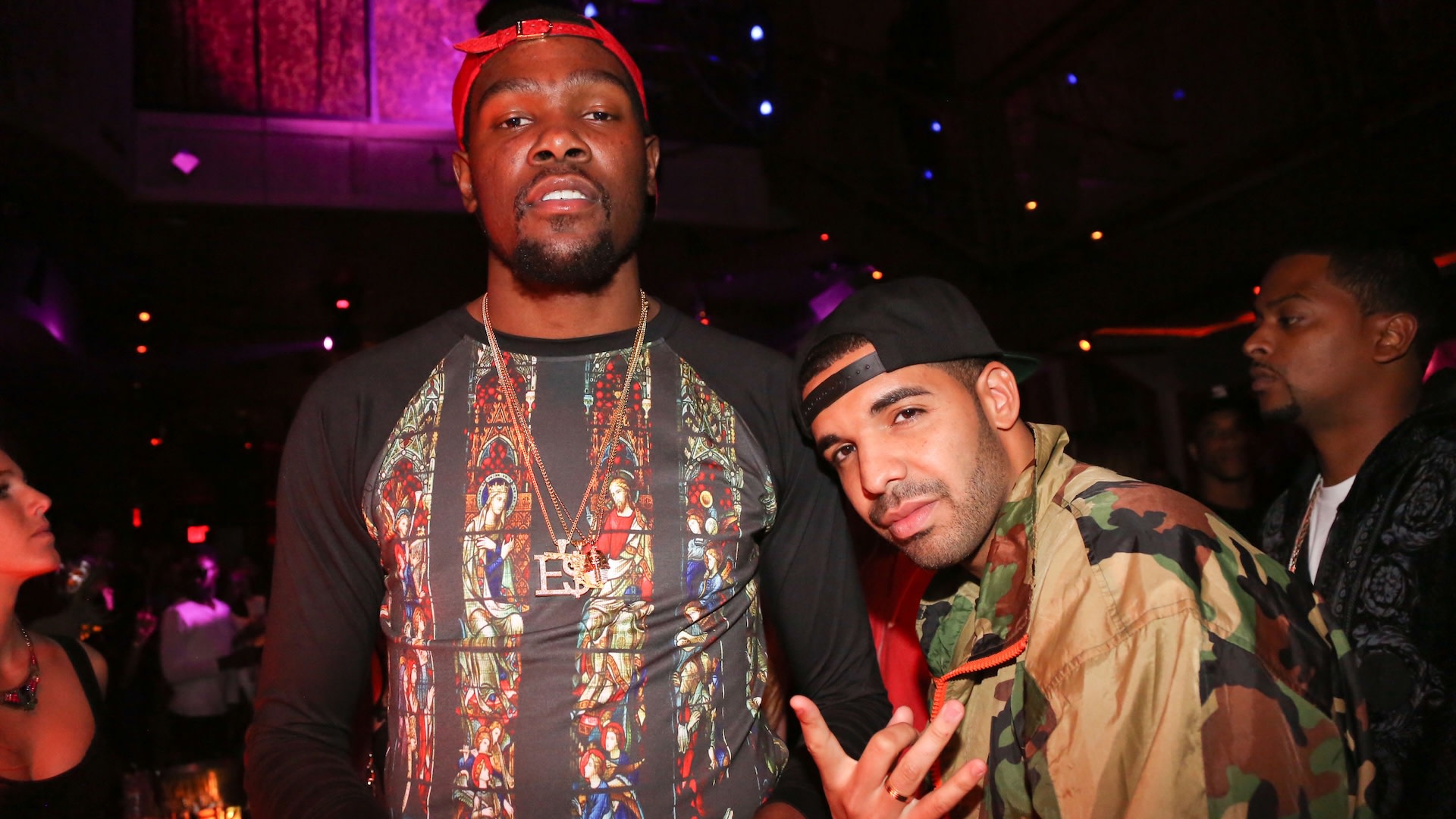 Kevin Durant and Drake are posing together in a dimly lit venue. Durant is wearing a patterned shirt and red cap, while Drake sports a camo jacket.