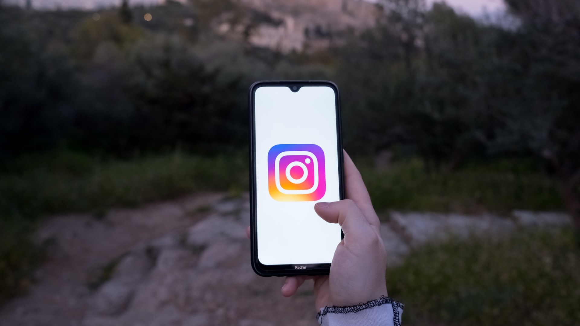 A person holding a smartphone displaying the Instagram logo, with an outdoor, blurred natural background.
