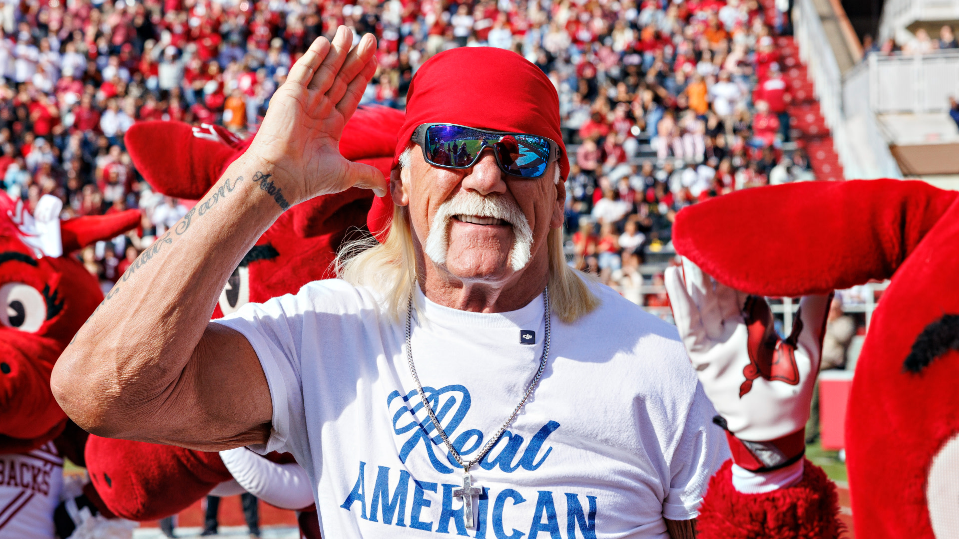 Hulk Hogan, wearing a red bandana and sunglasses, salutes at a sports event, surrounded by mascots and a cheering crowd.