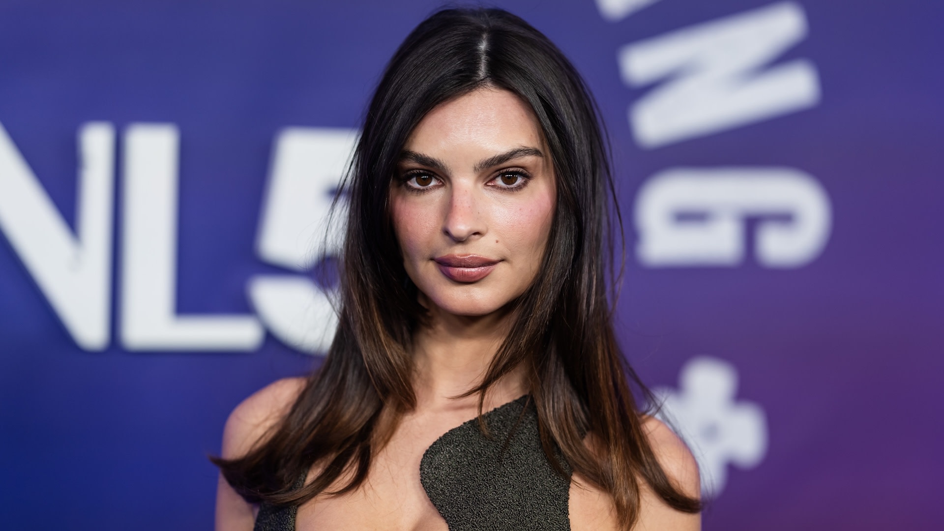 Emily Ratajkowski at an event, wearing a stylish dress with a backdrop featuring large text.