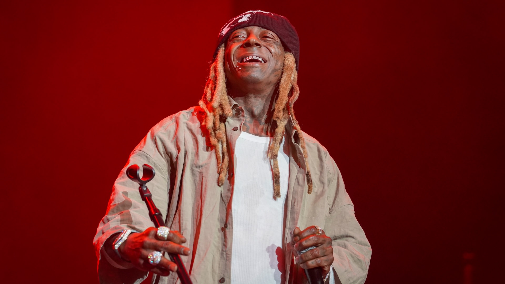 Lil Wayne performing on stage, wearing a beanie and casual outfit, smiling with a microphone in hand against a red-lit background.