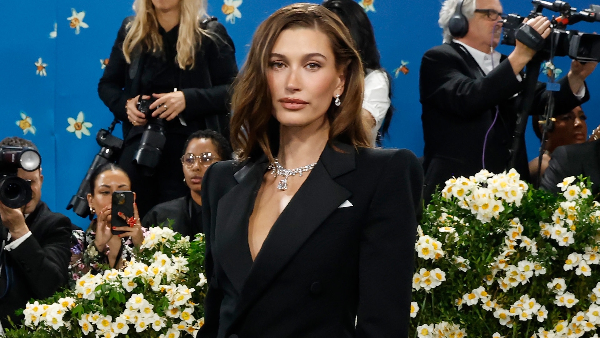 Hailey Bieber in a black suit, adorned with diamond jewelry, stands in front of a floral backdrop at a formal event.