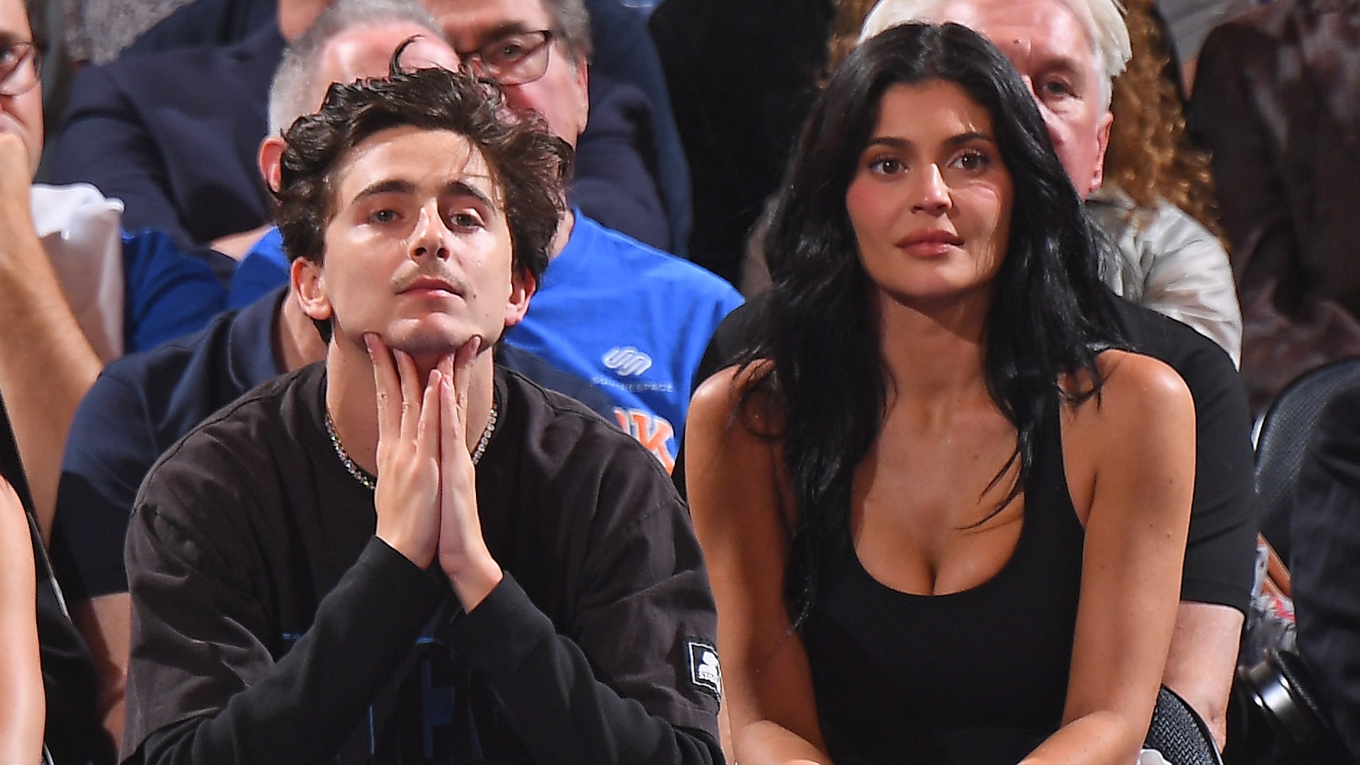 Timothée Chalamet and Kylie Jenner sitting courtside at a basketball game, surrounded by other spectators.