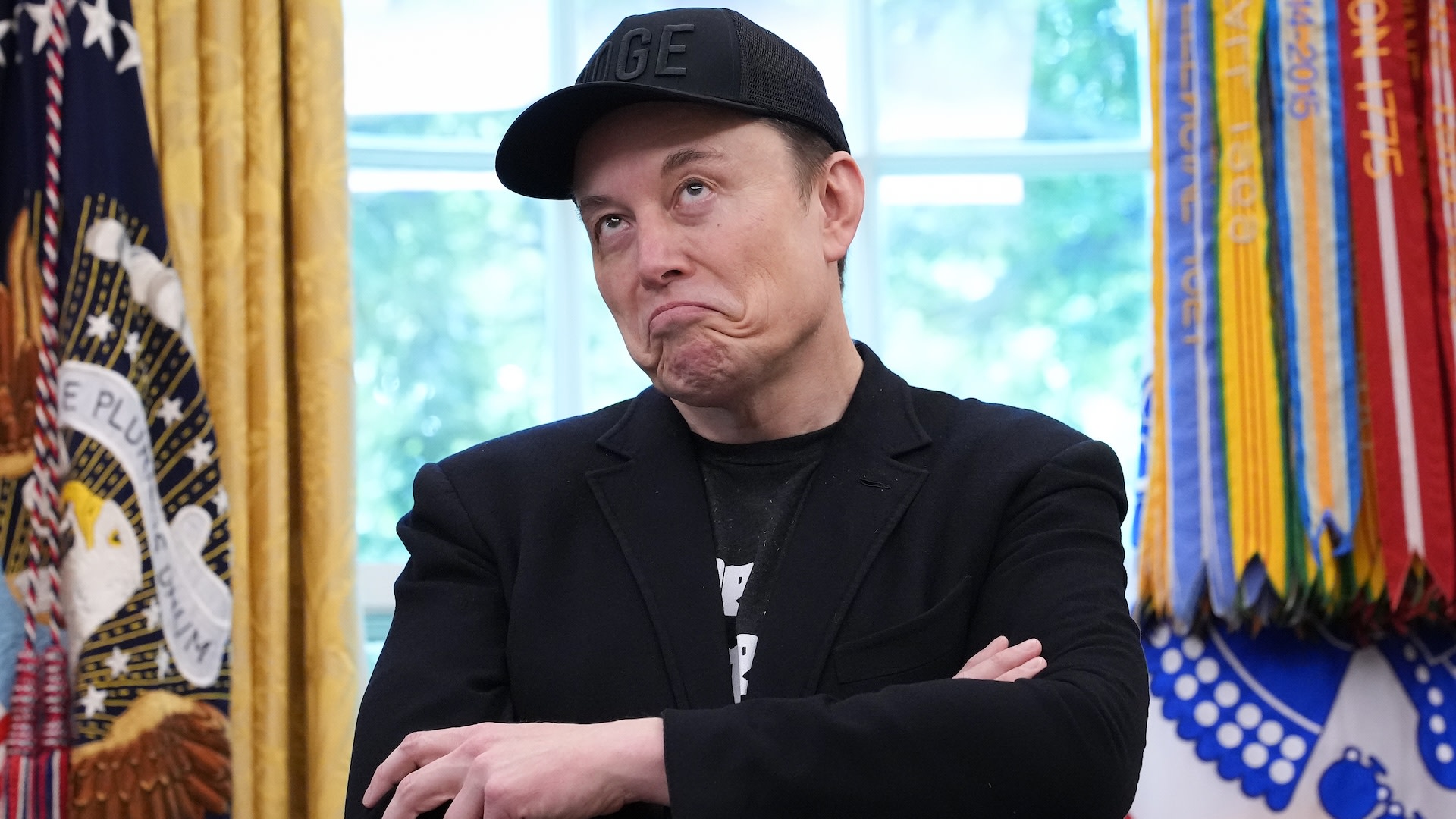 Elon Musk wearing a black cap and jacket, making a playful expression in a room with flags in the background.
