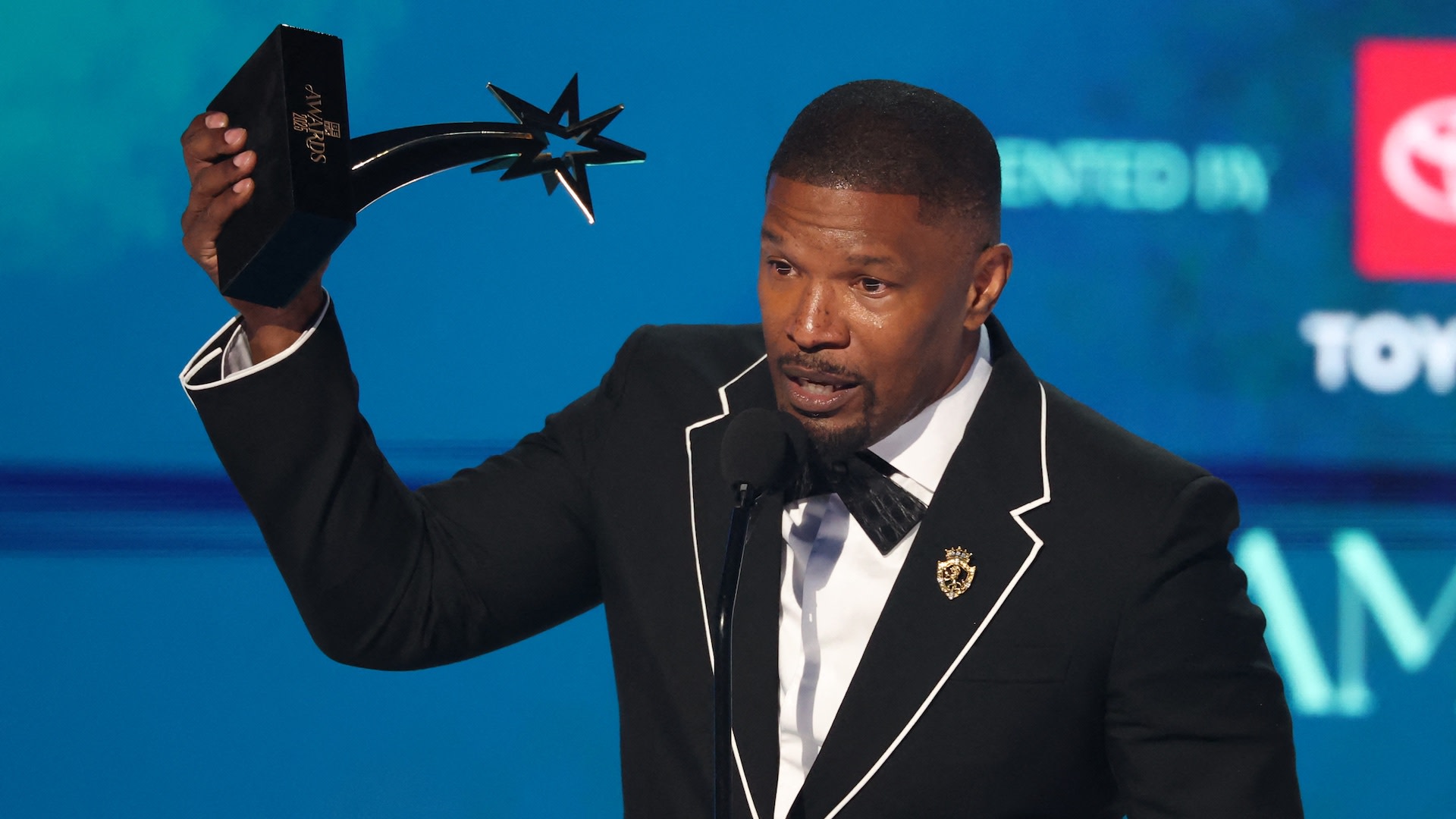 Jamie Foxx holding an award on stage, wearing a black suit with white trim and a bow tie, speaking into a microphone.
