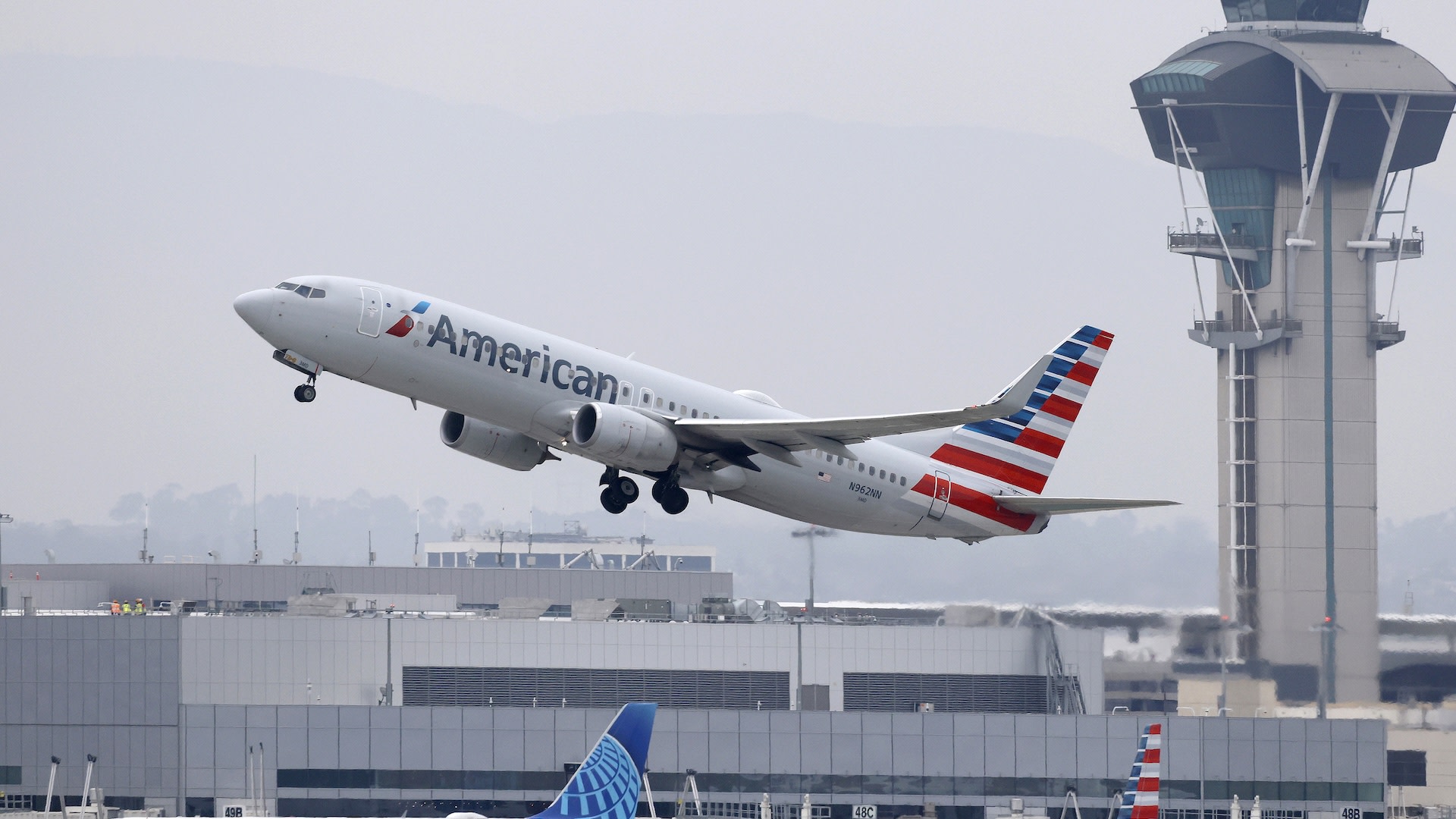 An American Airlines plane taking off from an airport, with a control tower and terminal buildings in the background.