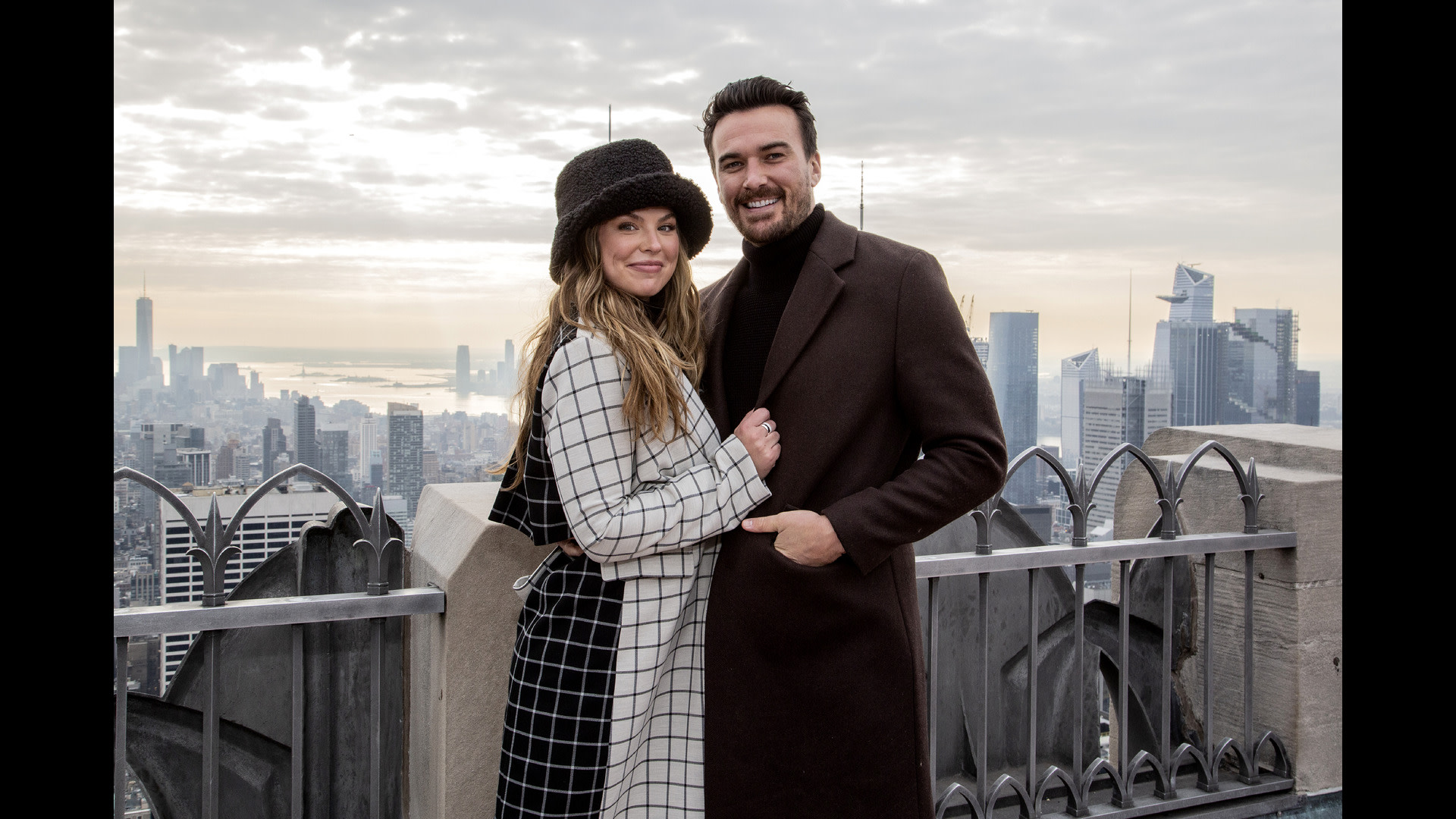 A couple poses on a rooftop with a cityscape background. The woman wears a checkered coat and hat, while the man is in a dark coat.