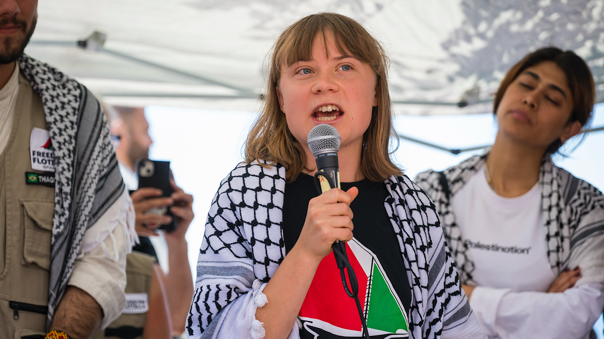 Greta Thunberg speaking into a microphone, wearing a black shirt and a keffiyeh. Others stand nearby, one with eyes closed.