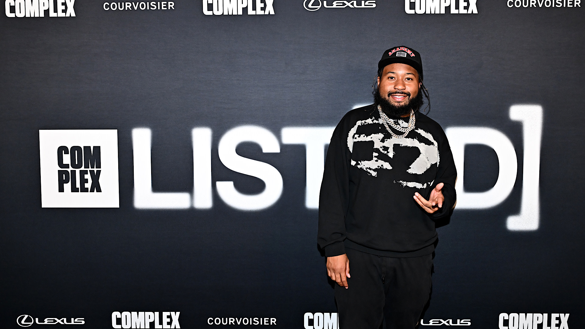 A man in a black outfit and cap stands smiling in front of a backdrop with "Complex Listed" and sponsor logos.