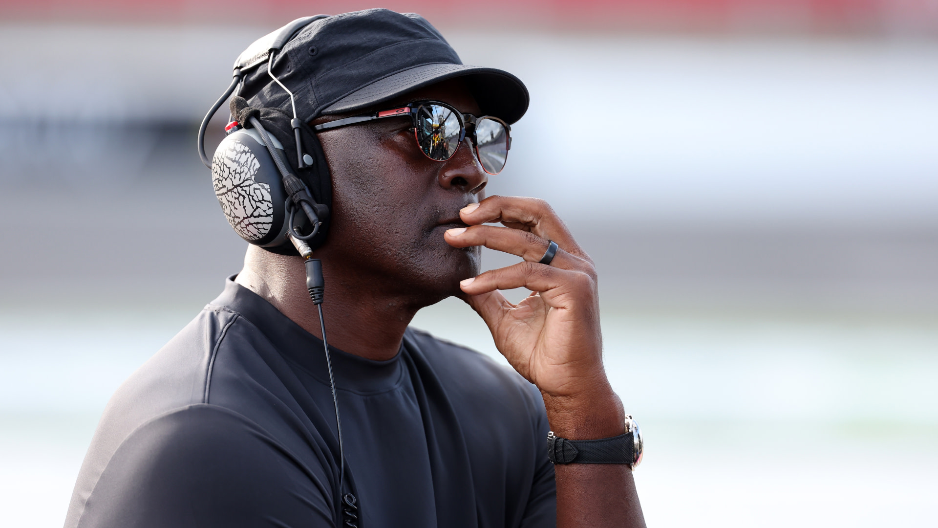 Michael Jordan wearing a black cap, sunglasses, and headphones, thoughtfully resting his hand on his chin.